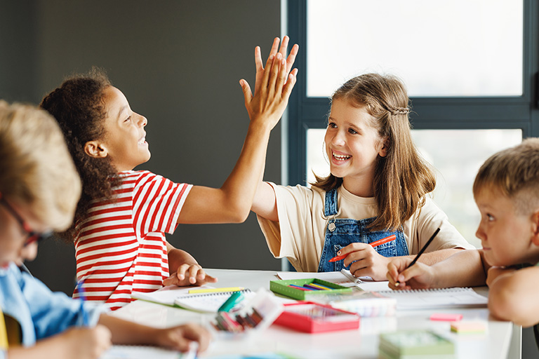 Two young female students, sitting at a table, high-fiving each other in a classroom setting, while two other student work.
