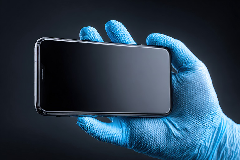 A hand covered in a blue latex glove hold a smartphone that is not powered on.