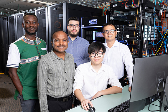 Five researchers stand together in a server room beside a desktop computer, with server racks, networking equipment, and cabling visible in the background.
